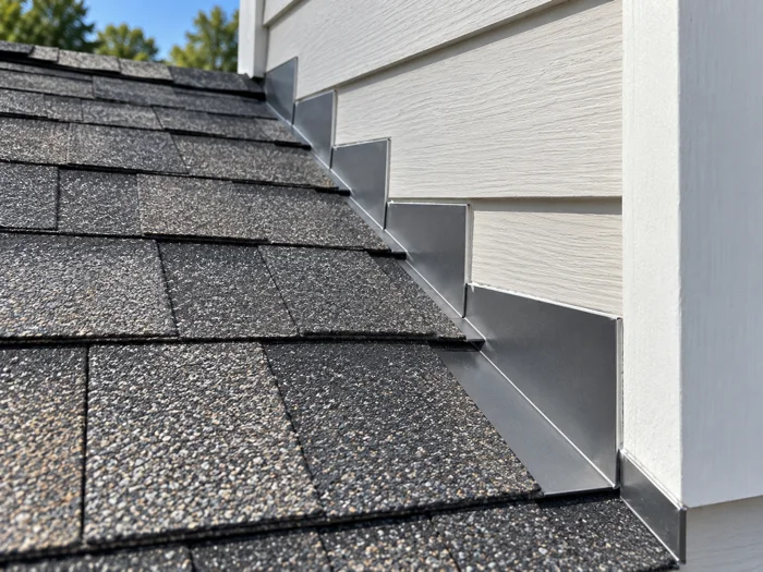 A close daylight detail of new architectural shingles meeting fresh step flashing at a wall, the lines straight, no people